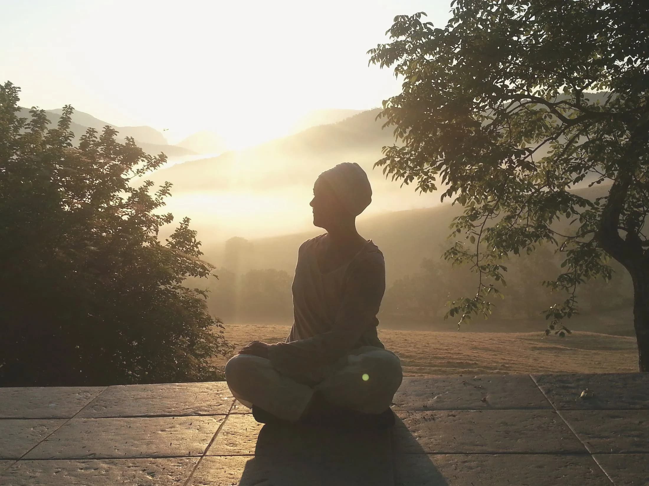 Amrita istuu meditaatiossa kivitasanteella auringonnousun aikaan, taustalla usvaiset kukkulat.